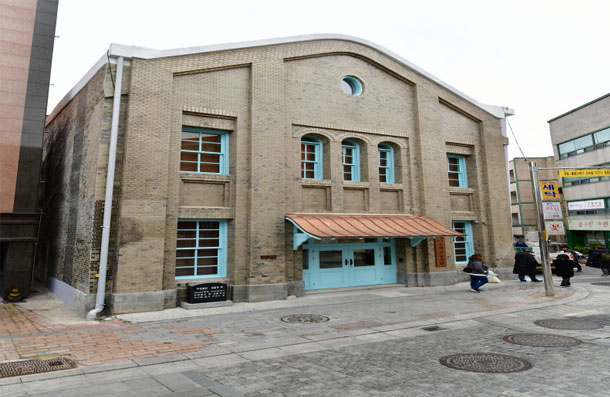 Front view of the two-story beige brick building of Former Bukukwon, showing its blue door and copper canopy