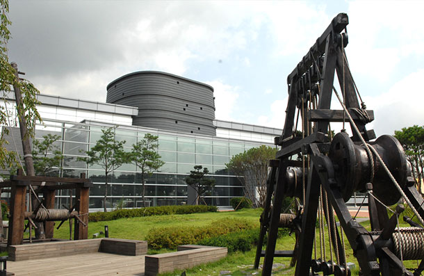 A replica of the geojunggi crane displayed in the outdoor garden of Suwon Hwaseong Museum, with the glass-walled building behind it